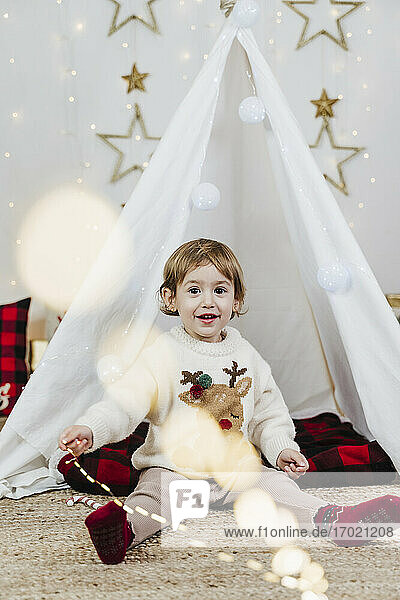 Niedliches kleines Mädchen  das beleuchtete Weihnachtslichter hält  während es in einem Zelt zu Hause sitzt