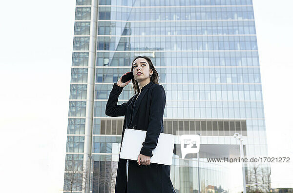 Young businesswoman with laptop talking on mobile phone while standing in city
