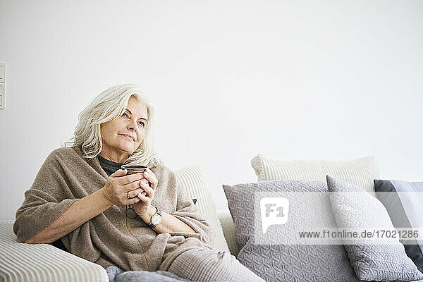 Nachdenkliche Frau im Ruhestand hält eine Kaffeetasse  während sie auf dem Sofa vor einer weißen Wand in einer Wohnung sitzt