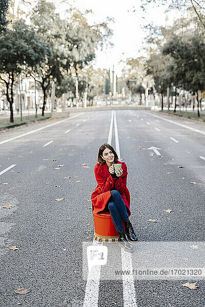 Junge Frau in Winterjacke trinkt Kaffee  während sie auf einem Sitz an der Straße sitzt und Musik hört