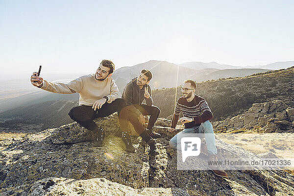 Young man taking selfie with friends while sitting on mountain during sunny day