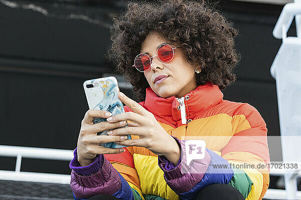 Curly hair woman wearing sunglasses and warm jacket using mobile phone while sitting outdoors
