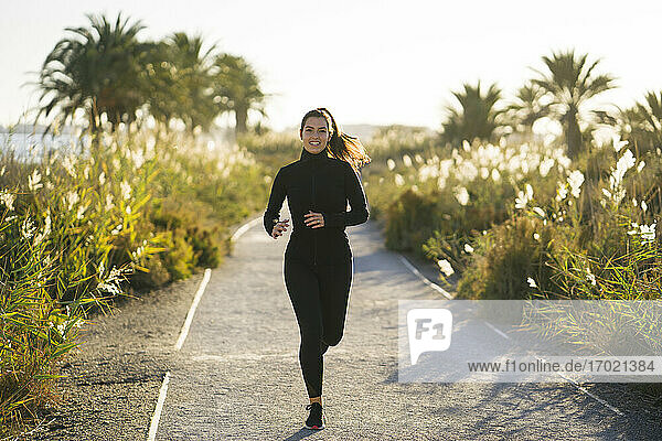 Lächelnde junge Frau  die bei Sonnenuntergang auf einem Fußweg inmitten von Pflanzen läuft