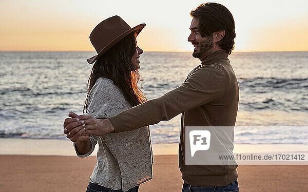 Smiling couple holding hands while dancing at beach during sunrise