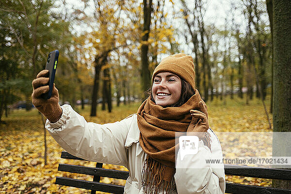 Glückliche junge Frau  die ein Selfie macht  während sie auf einer Bank im Herbstpark sitzt