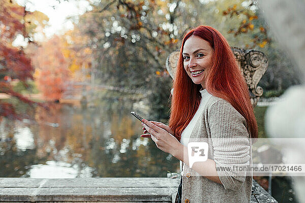 Lächelnde schöne Frau  die ein Mobiltelefon benutzt  während sie am See im Park steht