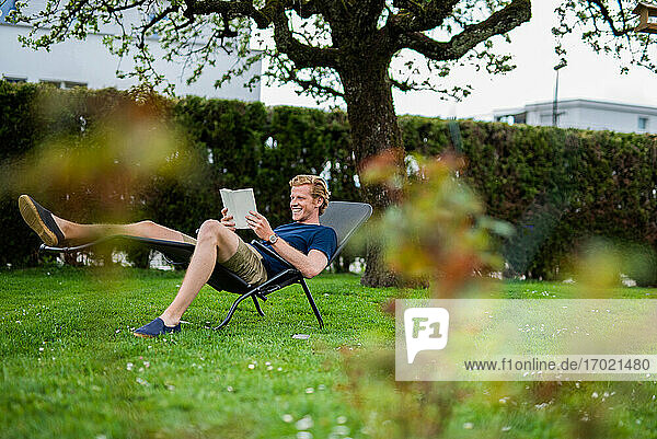 Smiling man reading book while sitting on chair in back yard