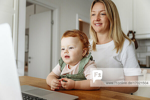 Woman with baby daughter using laptop at kitchen table