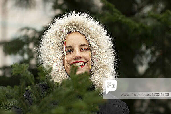 Lächelnde junge Frau in warmer Kleidung  die in der Weihnachtszeit vor einem Baum träumt