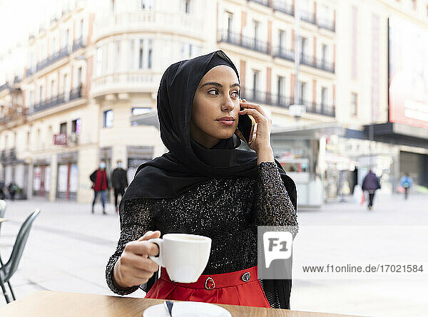 Porträt einer jungen schönen Frau mit schwarzem Hidschab  die in einem Straßencafé ein Smartphone benutzt