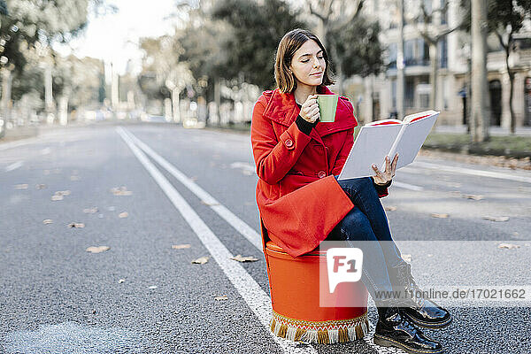 Modische junge Frau mit Kaffeetasse  die ein Buch liest  während sie auf einem Sitz an der Straße sitzt