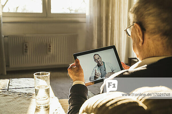 Männlicher Arzt berät einen älteren Mann per Videoanruf über ein digitales Tablet im Wohnzimmer