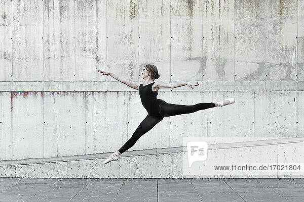 Ballerina im schwarzen Trikot springt vor einer Betonwand