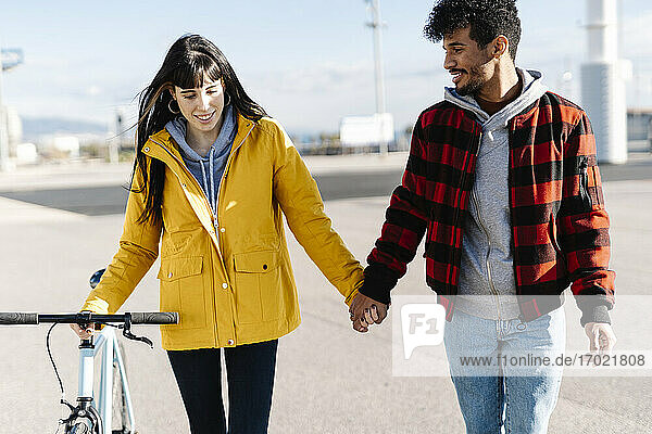 Smiling woman with bicycle holding hand of male friend while walking on footpath