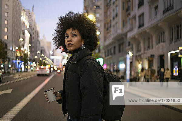 Junge Frau mit Einweg-Kaffeebecher beim Überqueren der Straße  während sie sich in der nächtlichen Stadt umsieht