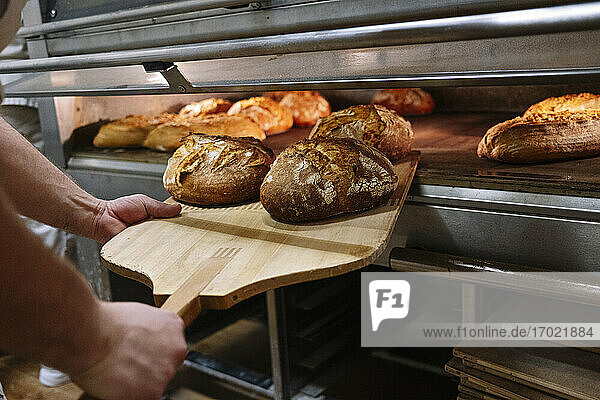 Männlicher Koch mit Pizzaschieber  der gebackenes Brot aus dem Ofen in einer Bäckerei nimmt