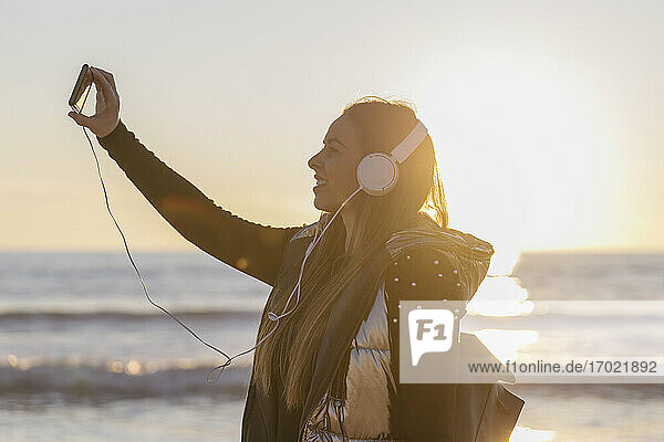 Schöne Frau  die selfie beim Hören von Musik gegen Meer während des Sonnenuntergangs