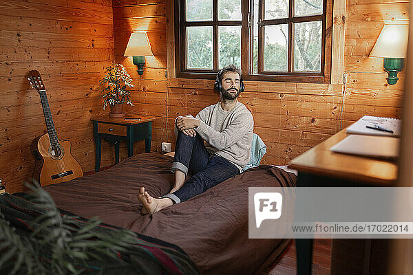 Young man with eyes closed music through headphones on bed in wooden cabin