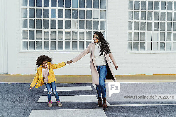 Fröhliche Mutter und Tochter halten sich an den Händen  während sie auf der Straße gegen ein Gebäude laufen