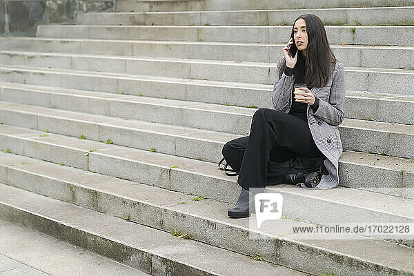 Unternehmerin mit wiederverwendbarem Kaffeebecher beim Telefonieren auf der Treppe sitzend