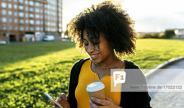 Lächelnde junge Frau mit lockigem Haar  die einen Kaffee in der Hand hält  während sie ein Mobiltelefon im Freien benutzt