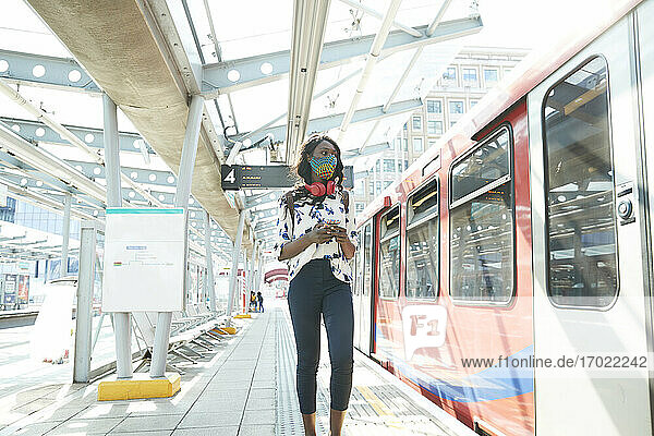 Geschäftsfrau mit Gesichtsmaske  die ein Mobiltelefon benutzt  während sie auf dem Bahnsteig steht
