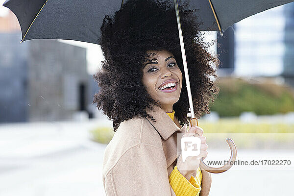 Lächelnde Frau mit Jacke  die einen Regenschirm hält  während sie im Freien steht