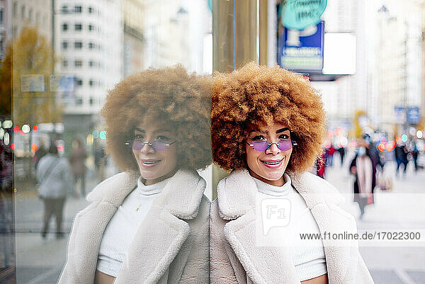 Lächelnde schöne Frau mit Afro-Haar lehnt an einer Glaswand in der Stadt