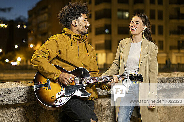Lächelnde Frau schaut auf einen Gitarre spielenden Mann  der sich auf eine Stützmauer stützt