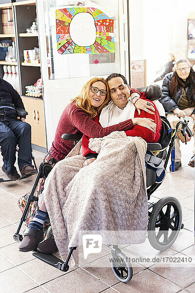 Weibliche Pflegekraft umarmt einen behinderten Mann  der in einem Pflegeheim im Rollstuhl sitzt