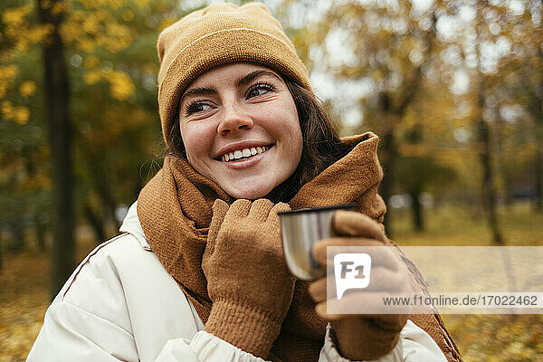 Happy young woman looking away while holding insulated cup in autumn park