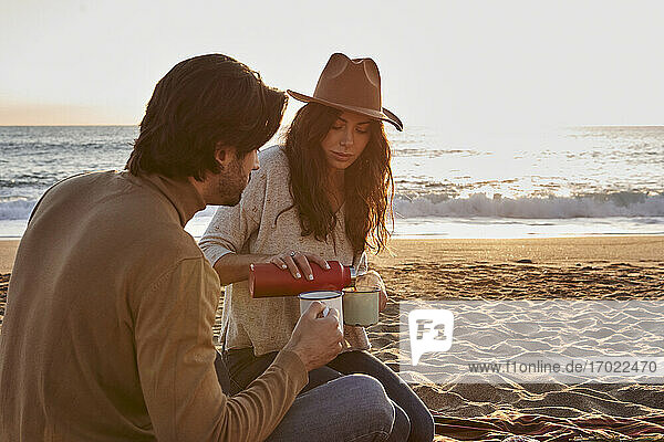 Frau mit Hut gießt Kaffee in eine Tasse und sitzt neben einem Mann am Strand