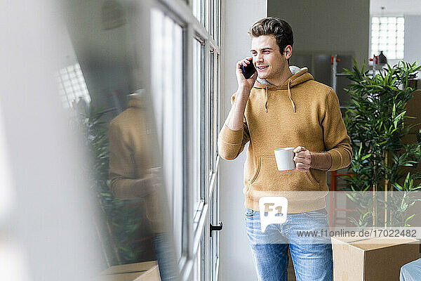 Lächelnder Mann telefoniert und hält eine Kaffeetasse am Fenster seines neuen Hauses