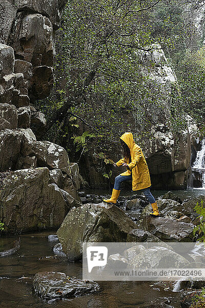Frau in gelbem Regenmantel klettert auf Felsen im Bach im Wald