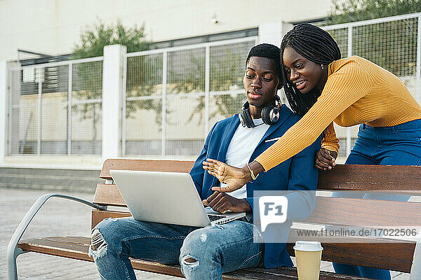 Lächelndes Teenager-Mädchen gestikuliert am Laptop  während es mit seinem Freund spricht