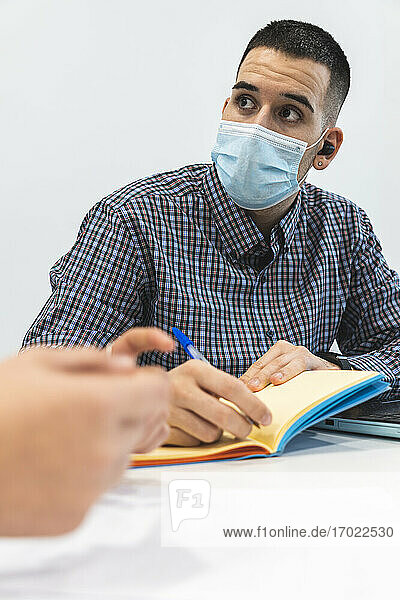 Geschäftsmann mit Gesichtsschutzmaske schreibt während einer Besprechung mit einem Kollegen im Büro in sein Tagebuch