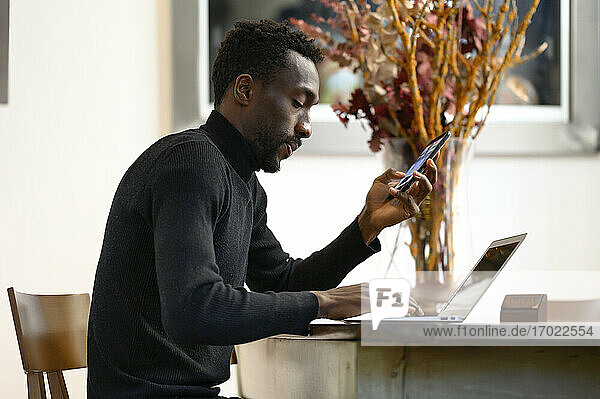 Junger Geschäftsmann mit Mobiltelefon  der in einem Café sitzt und am Laptop arbeitet