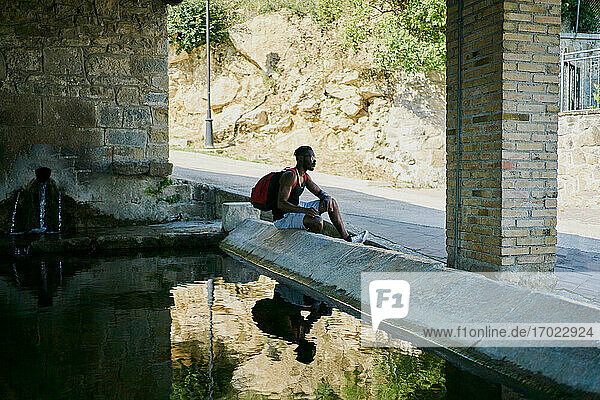 Junger Mann sitzt am Wasser und macht eine Pause beim Spaziergang