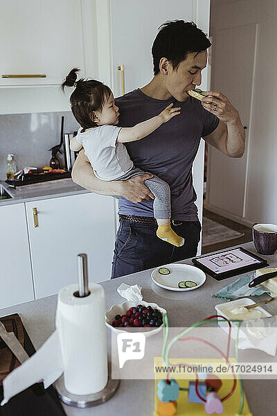 Father eating food while baby son looking at him in kitchen at home