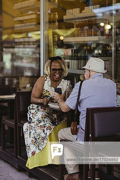 Senior woman laughing while man showing smart phone at sidewalk cafe
