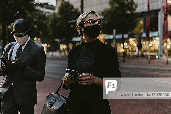 Weibliche Unternehmerin mit Smartphone schaut weg  während sie neben einem männlichen Kollegen steht  während COVID-19