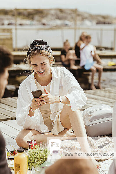 Lächelnde Frau mit Smartphone sitzen von Freund auf Steg während Picknick