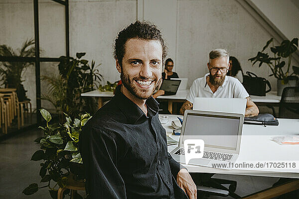 Porträt eines lächelnden männlichen Unternehmers  der im Büro am Laptop sitzt