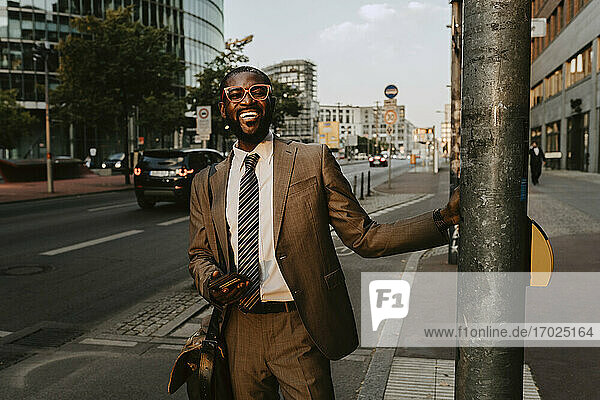 Smiling male entrepreneur standing by pole on footpath in city