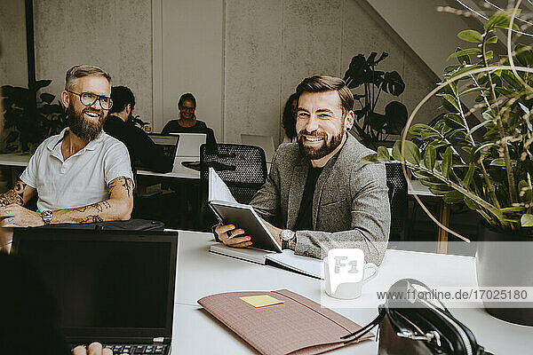 Lächelnde Geschäftsmann hält Tagebuch beim Sitzen mit männlichen Kollegen in Coworking Büro
