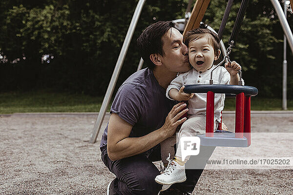 Lächelnder Vater küsst Sohn  während er auf einer Schaukel auf dem Spielplatz sitzt
