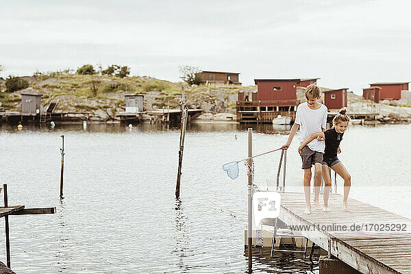 Siblings with fishing net walking on jetty at marina during vacation