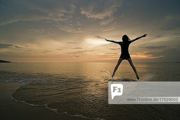 Eine junge Frau macht einen Sternsprung und genießt ihre Freiheit am Strand bei Sonnenuntergang.