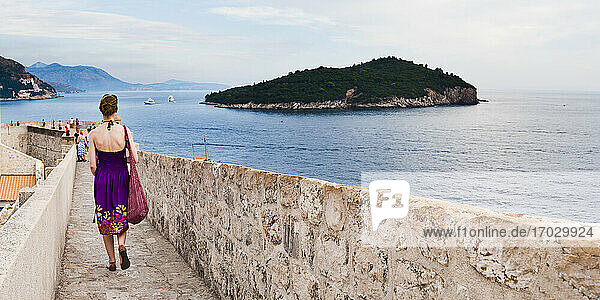 Panoramic photo of a tourist walking on Dubrovnik City Walls  Dubrovnik Old Town  Croatia. This is a panormic photo of a tourist walking on Dubrovnik City Walls  with Lokrum Island in the background. For tourists  Dubrovnik City Walls are one of the most popular things to do in Dubrovnik  as they provide incredible panoramic views over the whole of Dubrovnik Old Town and the Adriatic Coast  including Lokrum Island.