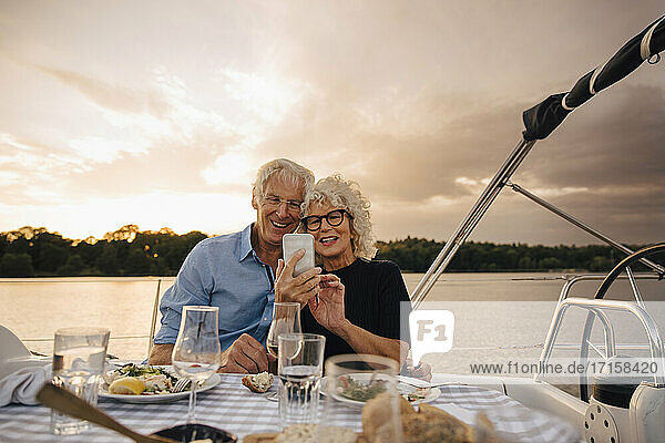Lächelndes heterosexuelles Paar nimmt Selfie durch Smartphone in Segelboot während Sonnenuntergang